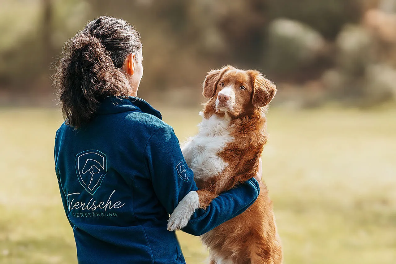 Frau in einer blauen Fleecejacke mit dem Logo von Tierische Verstärkung hält einen rot-weißen Retriever im Arm. Der Hund blickt aufmerksam zu ihr auf, während beide in einer ruhigen, natürlichen Umgebung stehen. Die Szene vermittelt Vertrauen, Verbundenheit und eine enge Mensch-Hund-Beziehung.