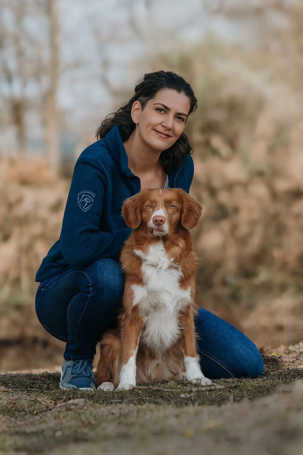 Die Inhaberin von Tierische Verstärkung kniet locker auf dem Boden, ihr rot-weißer Retriever sitzt vor ihr.