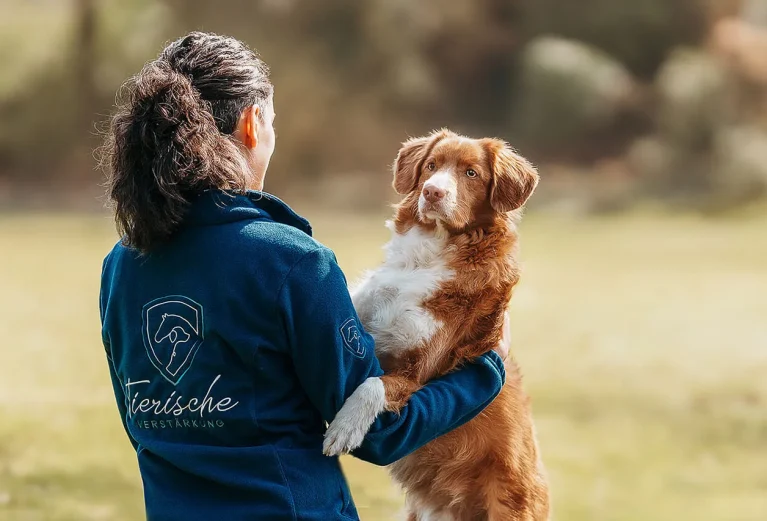 Frau in einer blauen Fleecejacke mit dem Logo von Tierische Verstärkung hält einen rot-weißen Retriever im Arm. Der Hund blickt aufmerksam zu ihr auf, während beide in einer ruhigen, natürlichen Umgebung stehen. Die Szene vermittelt Vertrauen, Verbundenheit und eine enge Mensch-Hund-Beziehung.