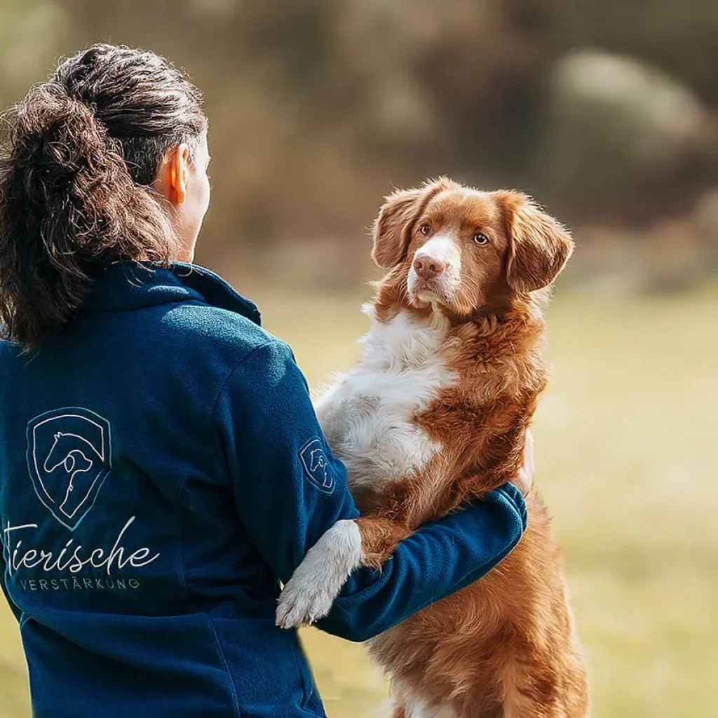 Frau in einer blauen Fleecejacke mit dem Logo von Tierische Verstärkung hält einen rot-weißen Retriever im Arm. Der Hund blickt aufmerksam zu ihr auf, während beide in einer ruhigen, natürlichen Umgebung stehen. Die Szene vermittelt Vertrauen, Verbundenheit und eine enge Mensch-Hund-Beziehung.