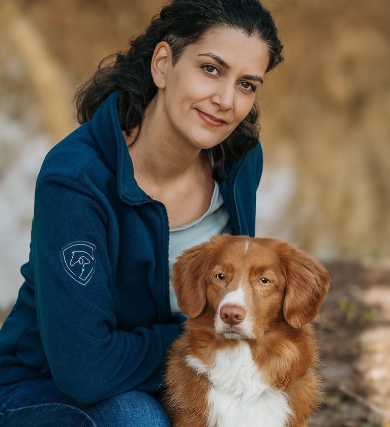 Die Inhaberin von Tierische Verstärkung kniet locker auf dem Boden, ihr rot-weißer Retriever sitzt vor ihr.